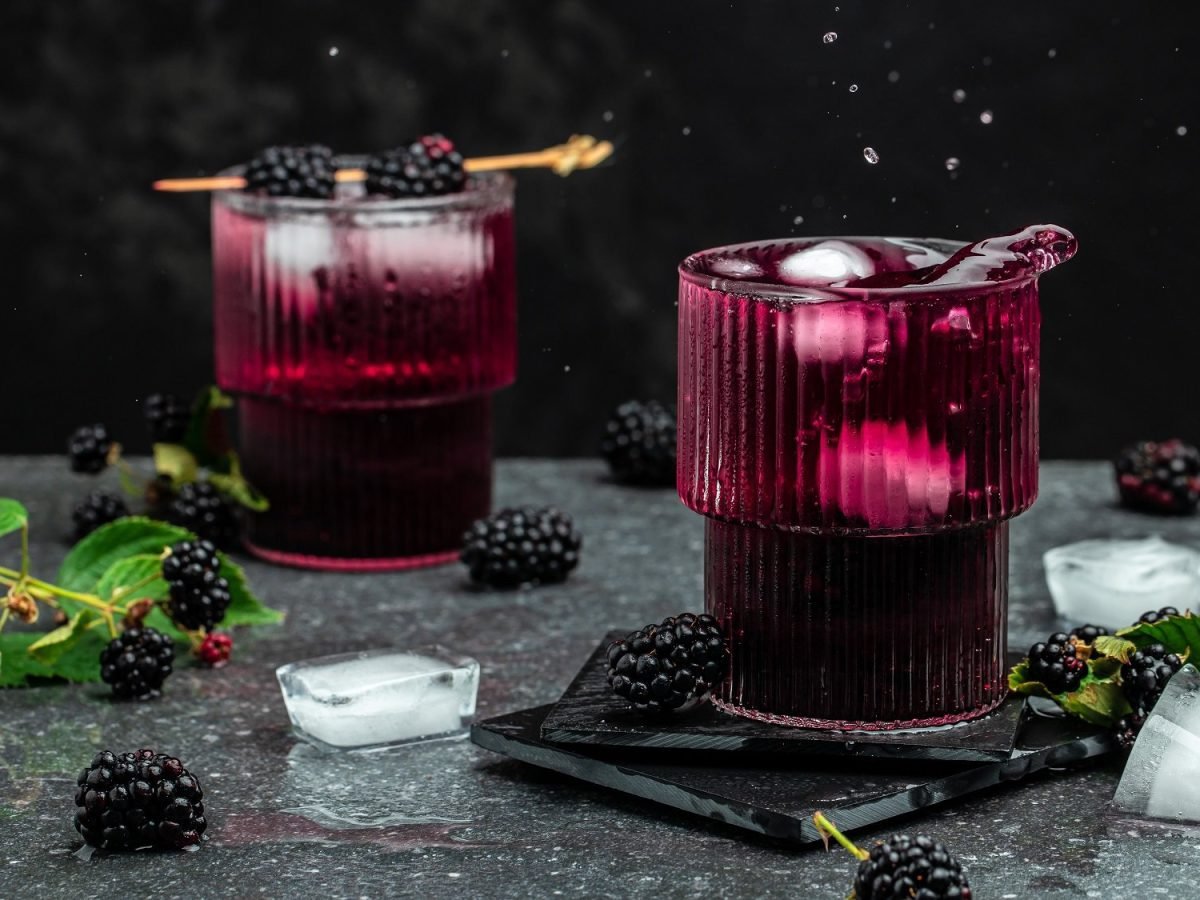 Zwei Gläser Brombeer-Mojito mit Eiswürfeln und Minze, dekoriert mit Brombeeren auf einem Zahnstocher, dunkle Umgebung, drumherum liegen Eiswürfel, Minze und Brombeeren.