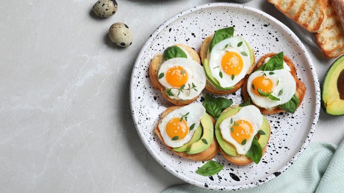 Mehrere Bruschetta mit Wachteleiern und Avocado auf einem dunkel-weiß gesprenkelten Teller von oben fotografiert. Daneben eine Avocado-Hälfte sowie Wachteleier.