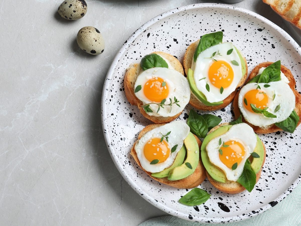 Mehrere Bruschetta mit Wachteleiern und Avocado auf einem dunkel-weiß gesprenkelten Teller von oben fotografiert. Daneben eine Avocado-Hälfte sowie Wachteleier.