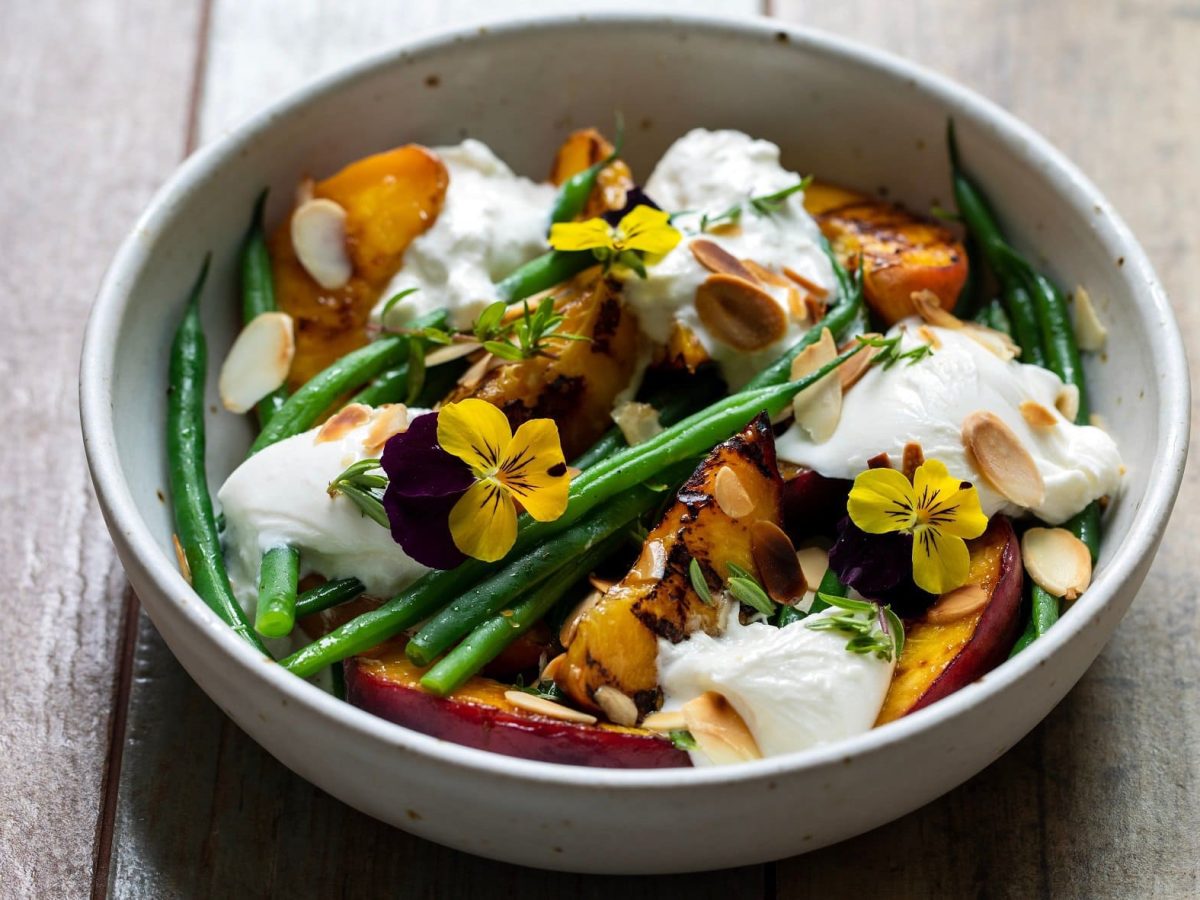 Schräge Draufsicht: Eine runde Bowl mit einem Burrata-Salat mit gegrillten Pfirsichen und grünen Bohnen auf einem Untergrund aus Holz.