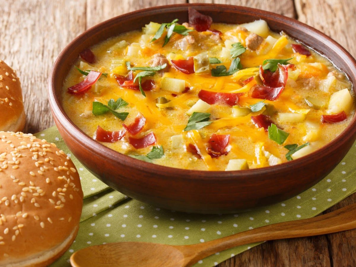 Cheeseburger Suppe mit Hackfleisch, Käse und Kartoffeln auf Serviette und Holztisch. Daneben zwei Burgerbrötchen. Unten im Bild ein Holzlöffel. Draufsicht.