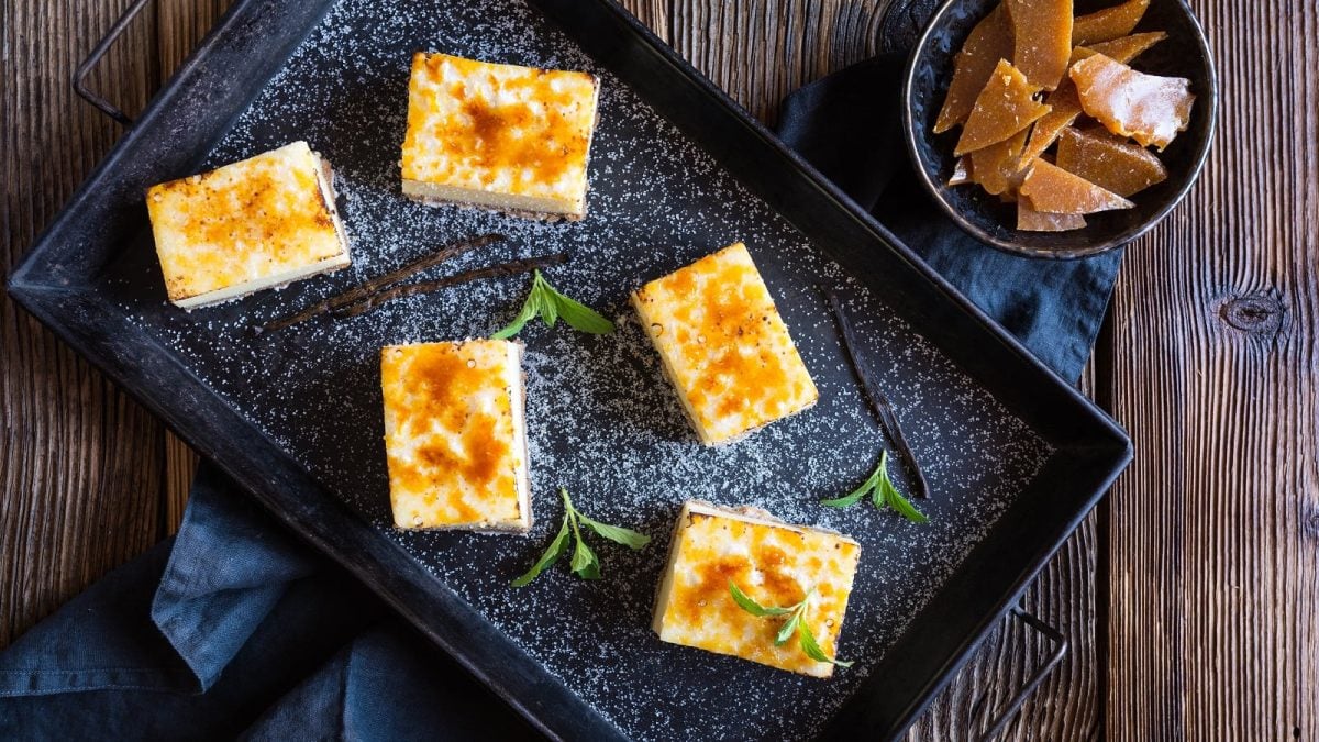 Eine schwarze Platte mit Crème Brûlée Cheesecake Bars auf einem Holztisch.