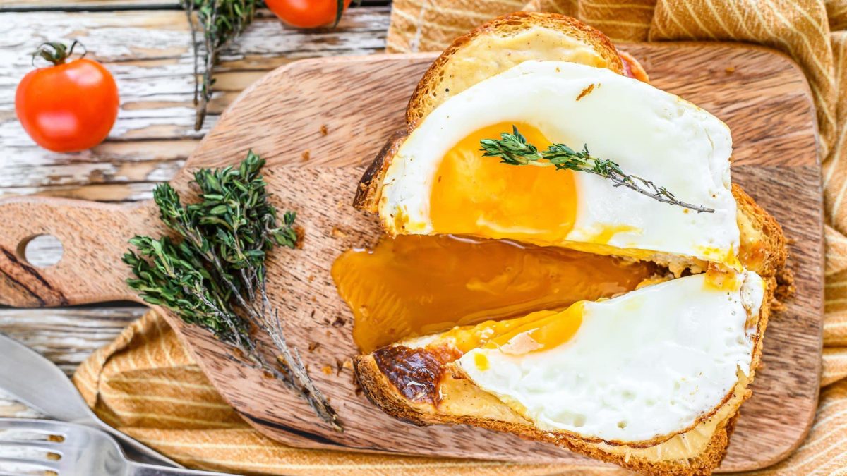 Croque Madame auf Holzbrett mit angeschnittenem Spiegelei. Im Bild frischer Thymian und kleine Tomaten. Draufsicht.