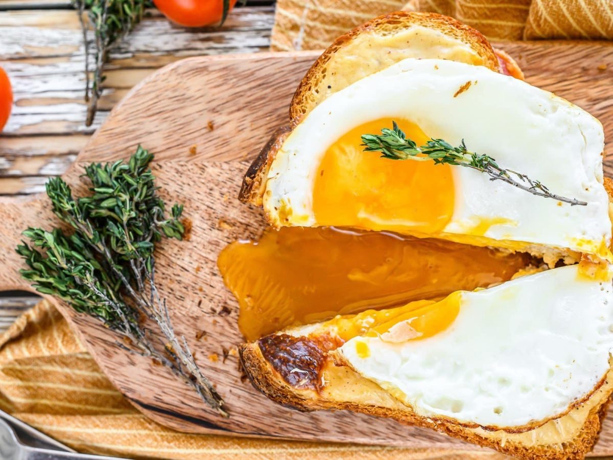 Croque Madame auf Holzbrett mit angeschnittenem Spiegelei. Im Bild frischer Thymian und kleine Tomaten. Draufsicht.