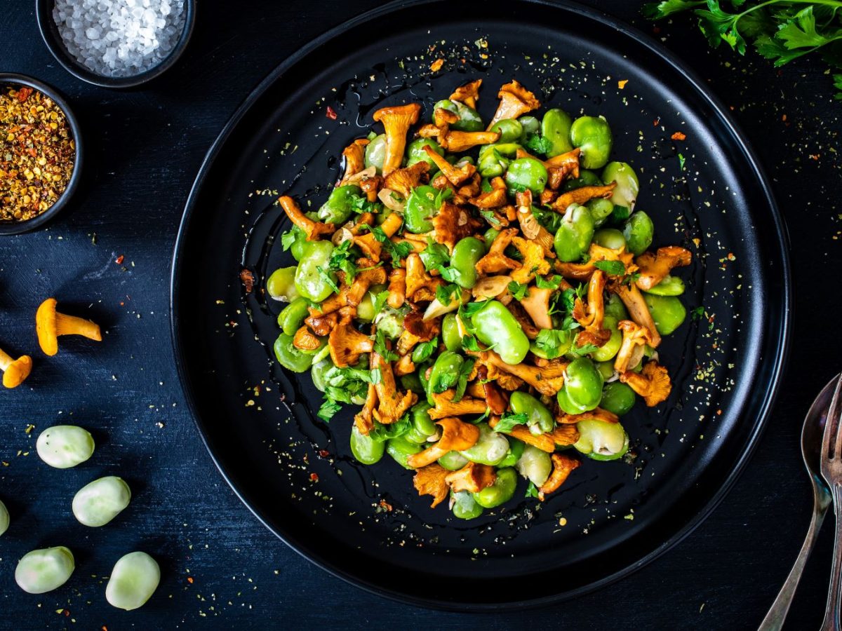 Edamame Salat mit sautierten Pfifferlingen in schwarzem Teller auf dunklem Untergrund. Im Bild rohe Bohnen, Pfifferlinge, Petersilie und, Besteck und Gewürze in schwarzen Schüsseln. Draufsicht