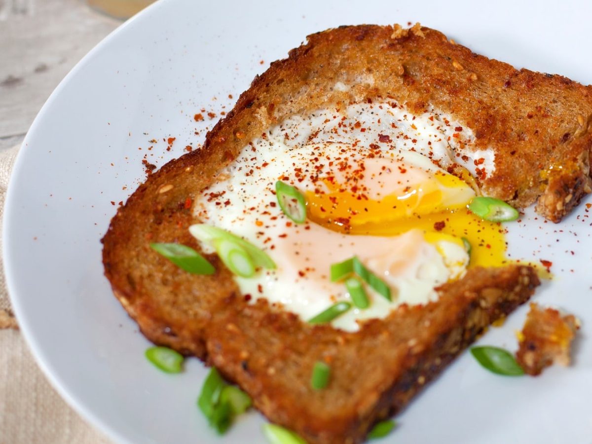 Eine Brotscheibe mit dem Egg in a Hole zusammen mit Chilipulver und Frühlingslauch in sehr naher Aufnahme.