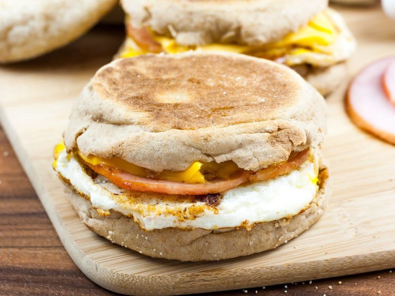 Egg McMuffin mit Bacon auf Holzplatte. Im Hintergrund Kochschinken und weitere englische Muffins auf Holztisch. Draufsicht.