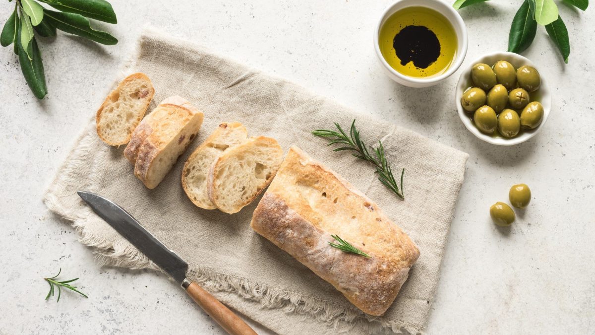 Einfaches Ciabatta Rezept ein angeschnittenes Ciabatta auf Tuch auf hellem Untergrund. Im Bild ein Messer mit Holzgriff, eine Schale mit Öl und Essig sowie eine mit grünen Oliven. Olivenzweige und Rosmarin am Bildrand. Draufsicht.