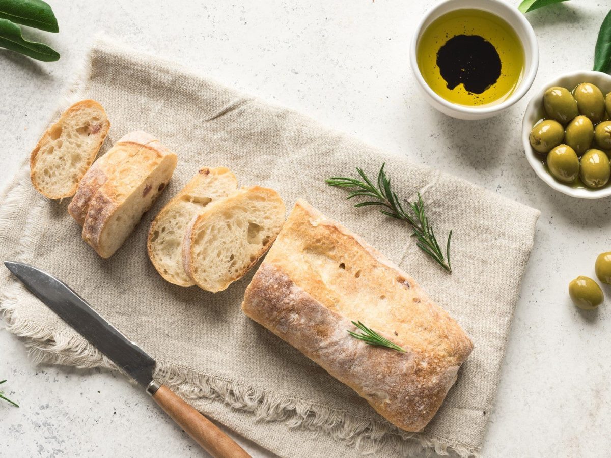 Einfaches Ciabatta Rezept ein angeschnittenes Ciabatta auf Tuch auf hellem Untergrund. Im Bild ein Messer mit Holzgriff, eine Schale mit Öl und Essig sowie eine mit grünen Oliven. Olivenzweige und Rosmarin am Bildrand. Draufsicht.