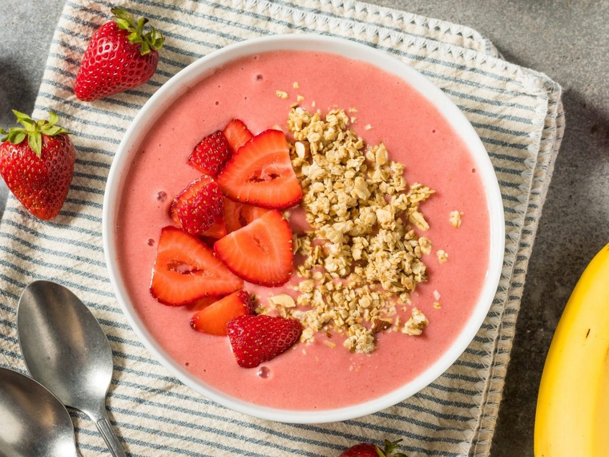 Eine weiße Schüssel mit der Erdbeer-Joghurt-Bowl auf einem grauen Küchentisch mit Erdbeeren und Banane.