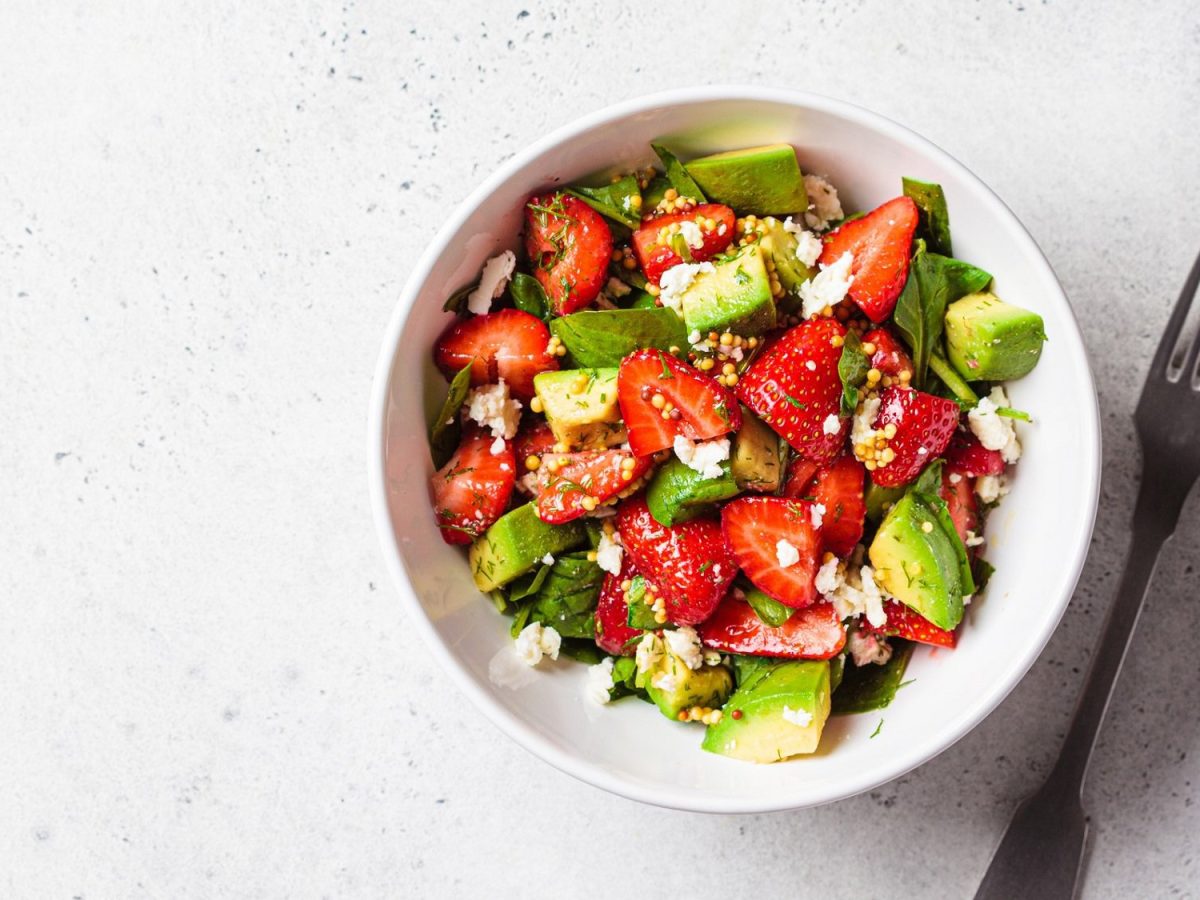 Eine Schale Erdbeer-Spinat-Salat mit Avocado & Feta in der Draufsicht auf hellem Untergrund, daneben eine Gabel.