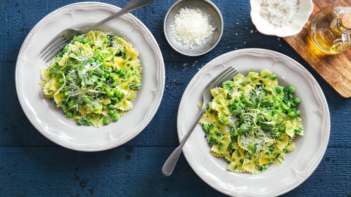 Farfalle mit Brokkoli-Minz-Pesto und Erbsen auf zwei Pastatellern auf blauem Untergrund. Im Bild ein Holzbrett mit einer Karaffe Olivenöl und zwei Schalen mit geriebenem Käse. Draufsicht.