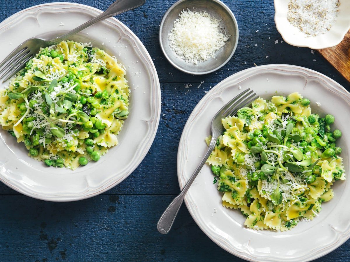 Farfalle mit Brokkoli-Minz-Pesto und Erbsen auf zwei Pastatellern auf blauem Untergrund. Im Bild ein Holzbrett mit einer Karaffe Olivenöl und zwei Schalen mit geriebenem Käse. Draufsicht.