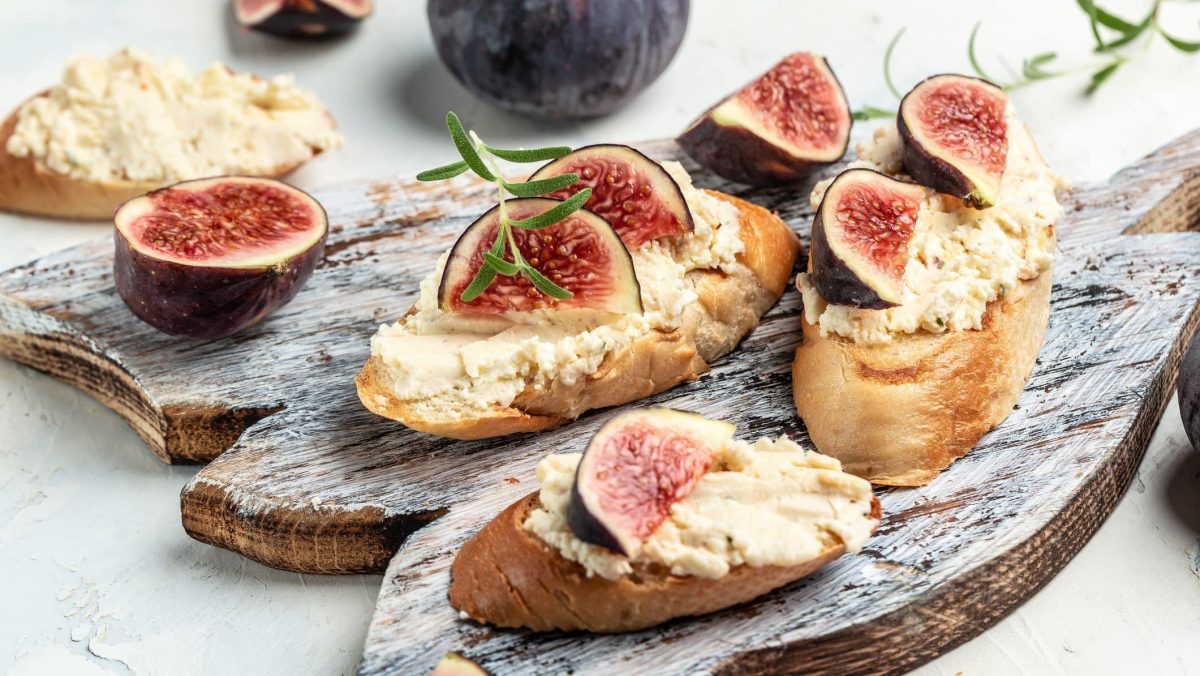 Feigenaufstrich mit Ziegenfrischkäse und frischen Feigen auf Brotscheiben mit frischem Rosmarin auf Holzbrett und hellem Untergrund. Im Bild Feigen, Rosmarin und weitere Brotscheiben. Frontalansicht.