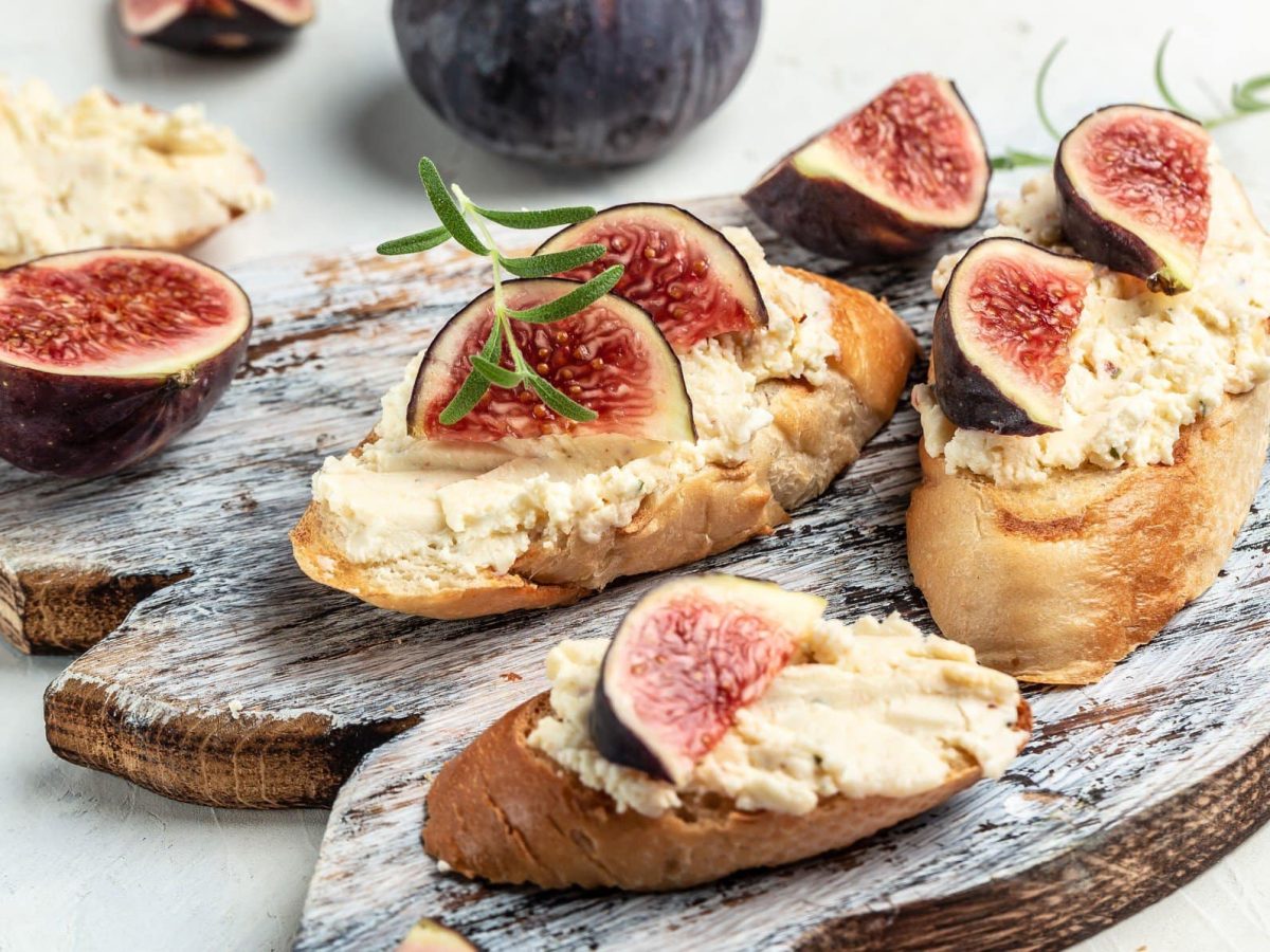 Feigenaufstrich mit Ziegenfrischkäse und frischen Feigen auf Brotscheiben mit frischem Rosmarin auf Holzbrett und hellem Untergrund. Im Bild Feigen, Rosmarin und weitere Brotscheiben. Frontalansicht.