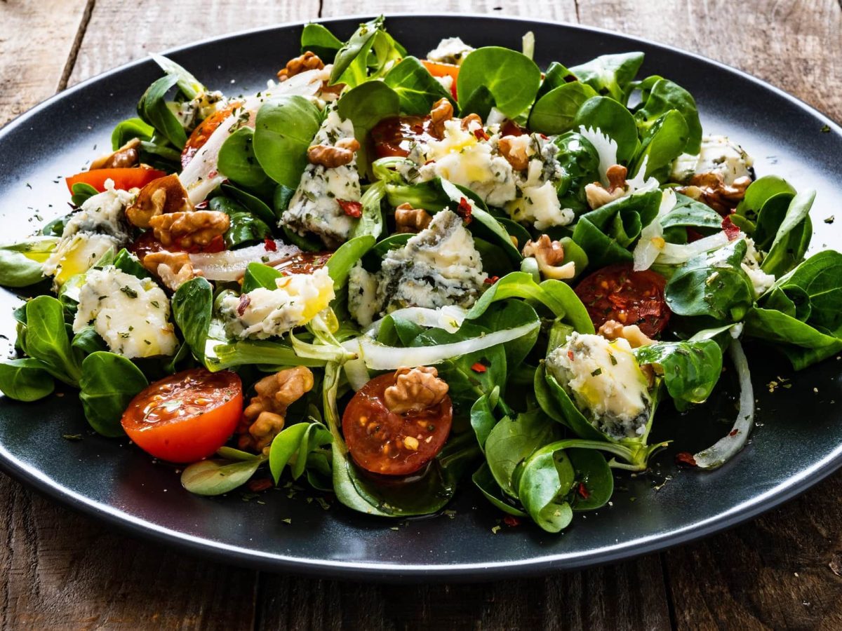 Schräge Draufsicht: ein dunkler Teller mit Feldsalat mit Roquefort, Tomaten, schwarzem Sesam und Walnüssen.