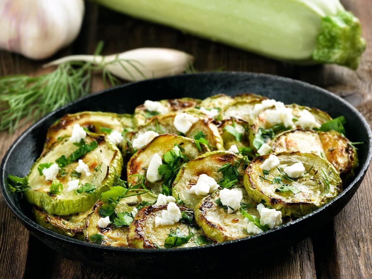 Schräge Draufsicht: Eine Pfanne frittierte Zucchini mit Feta auf einem Untergrund aus Holz.