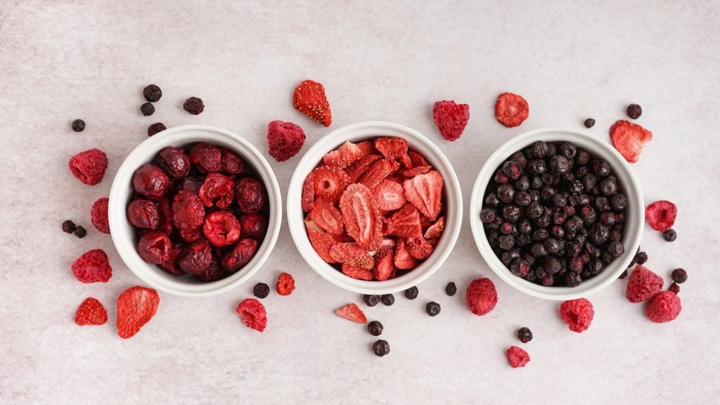 Gefriergetrocknete Kirschen, Erdbeeren und schwarze Beeren in weißen Schalen für selbstgemachtes Müsli, umgeben von verstreuten Beeren auf hellem Grund.