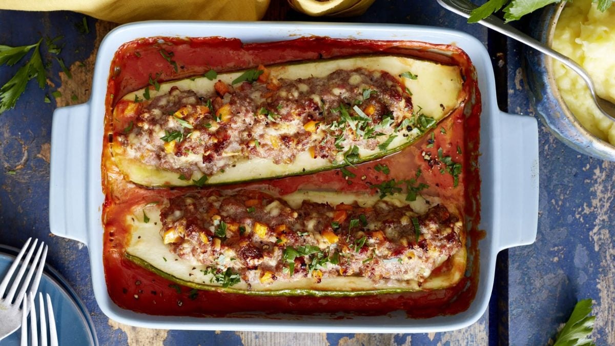 Gefüllte Schmorgurken mit Mett und Käse in Tomatensauce in Auflaufform auf blauen Untergrund. Draufsicht.