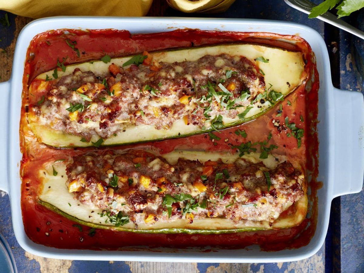 Gefüllte Schmorgurken mit Mett und Käse in Tomatensauce in Auflaufform auf blauen Untergrund. Draufsicht.