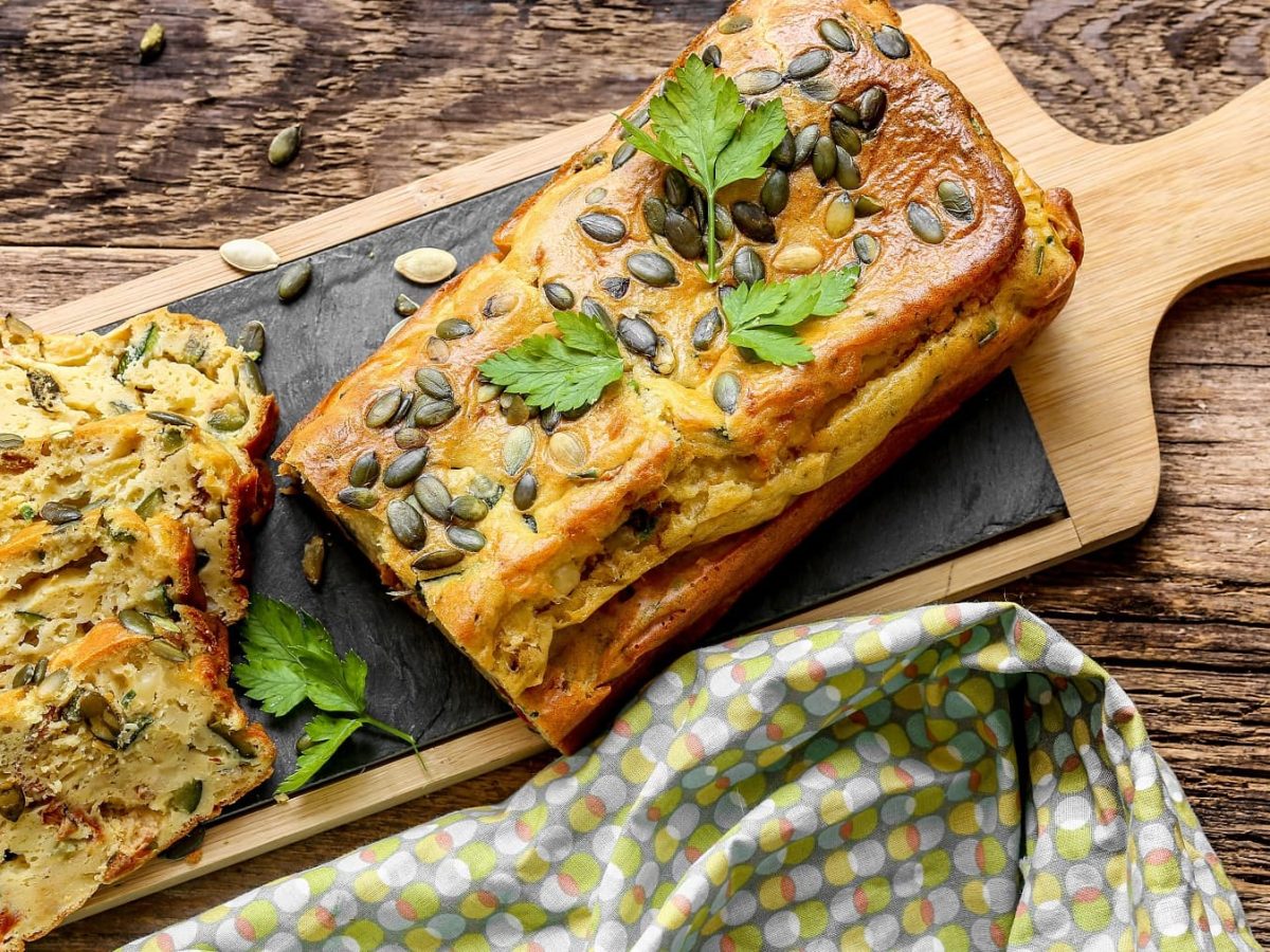 Ein Gemüsebrot in Kastenform auf einem Holzschneidebrett und dunklem Holztisch mit einem grünlichen Tuch von oben fotografiert.