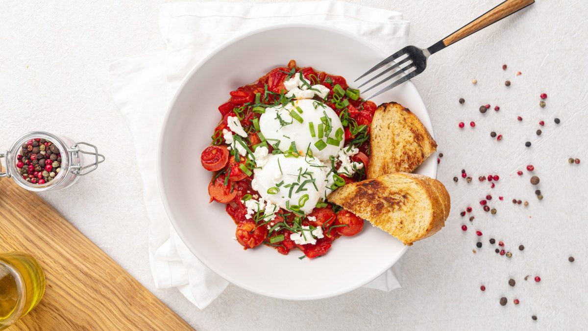 Draufsicht: weißer Untergrund, weiße Schüssel mit geschmorten Kirschtomaten mit pochiertem Ei. Daneben liegen Pfefferkörner verteilt und in einer kleinen Schüssel.