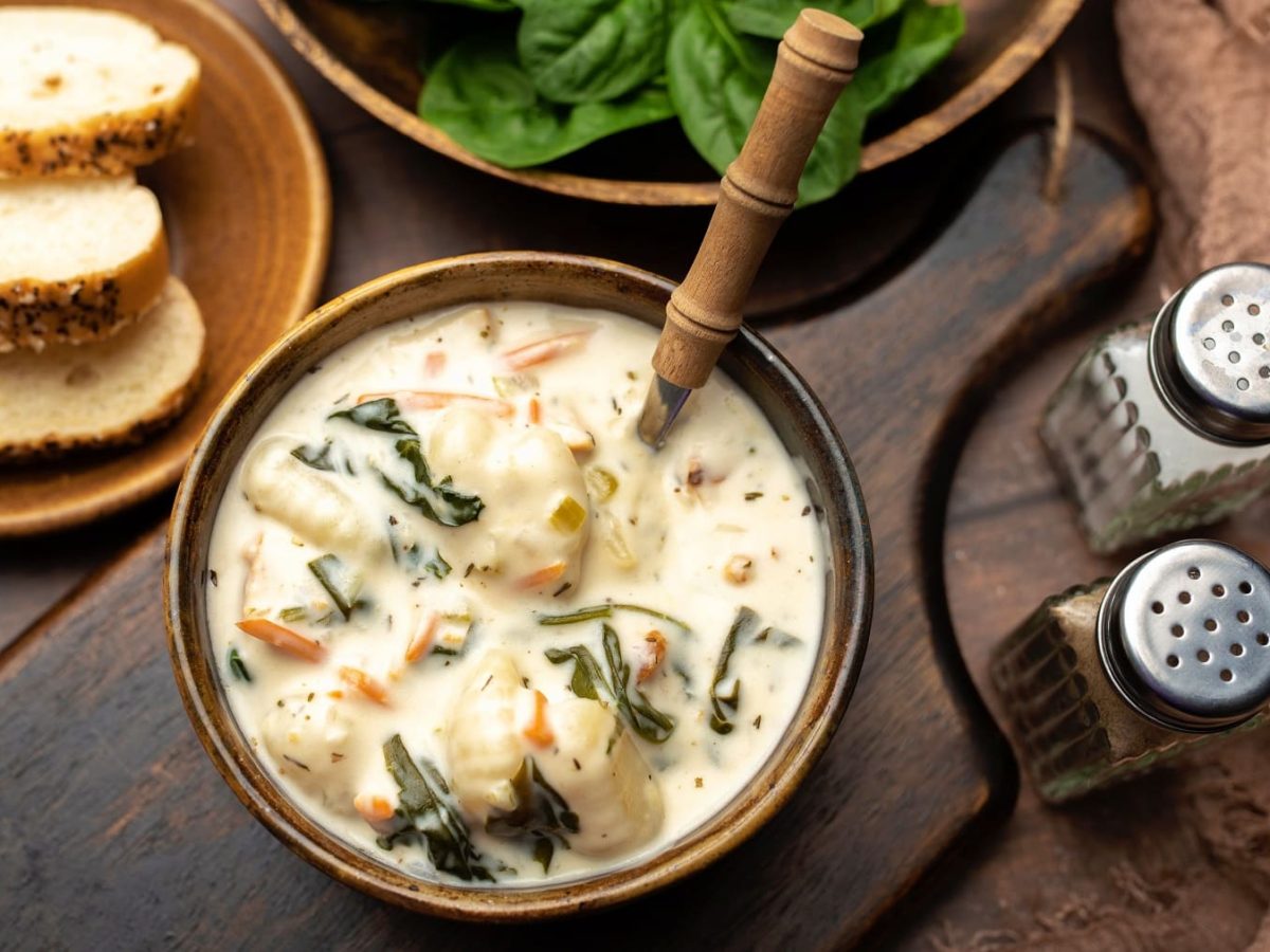 Eine braune Holzschüssel mit der Gnocchi-Suppe auf einem dunklen Holzbrett von oben fotografiert. Daneben Salz- und Pfefferstreuer sowie Brotscheiben und Babyspinat.