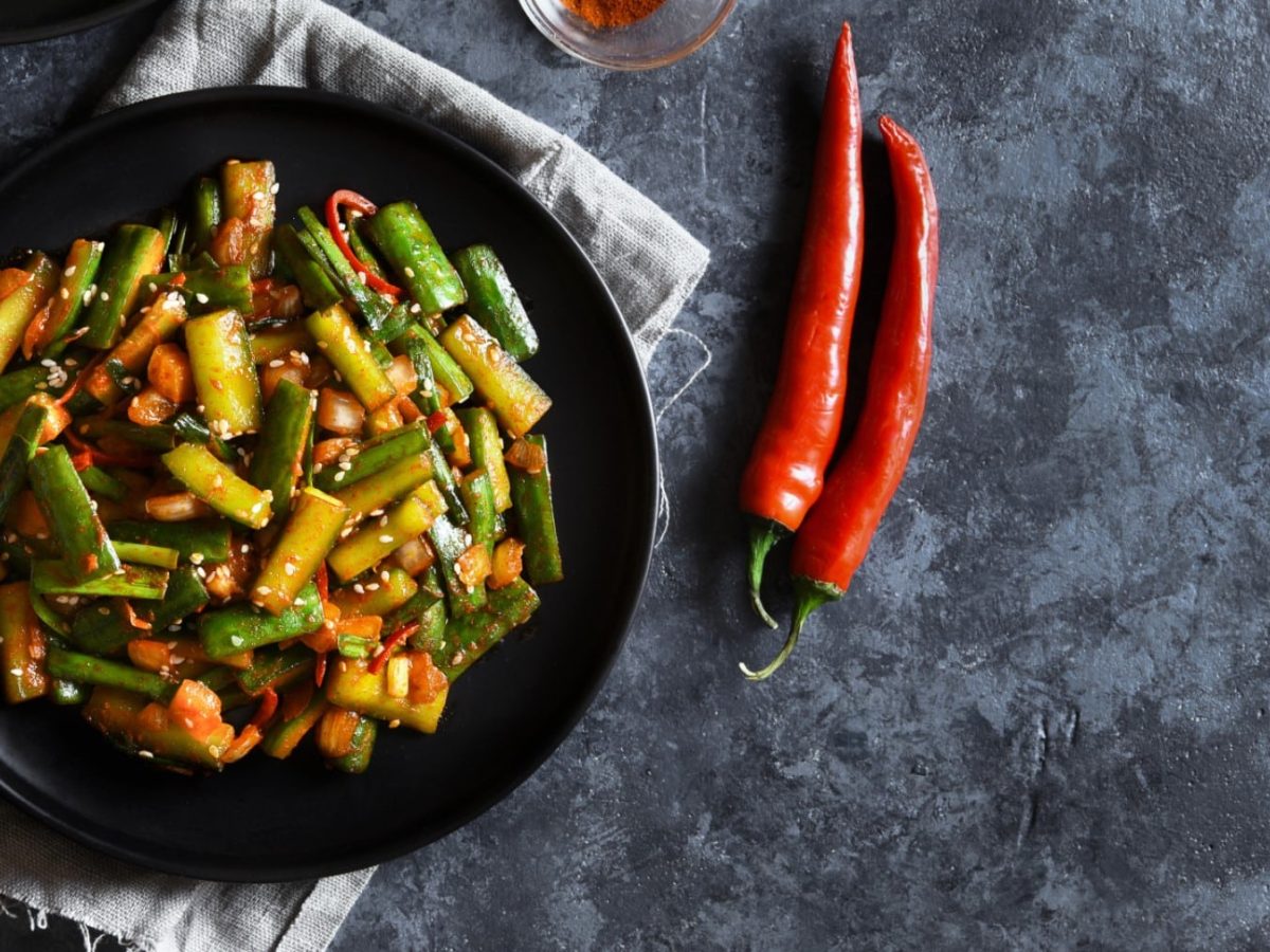 Gurken-Kimchi in einer schwarzen Bowl auf einem grauen Geschirrtuch auf einem dunkelgrauen Hintergrund. Rechts davon zwei rote Chilischoten.