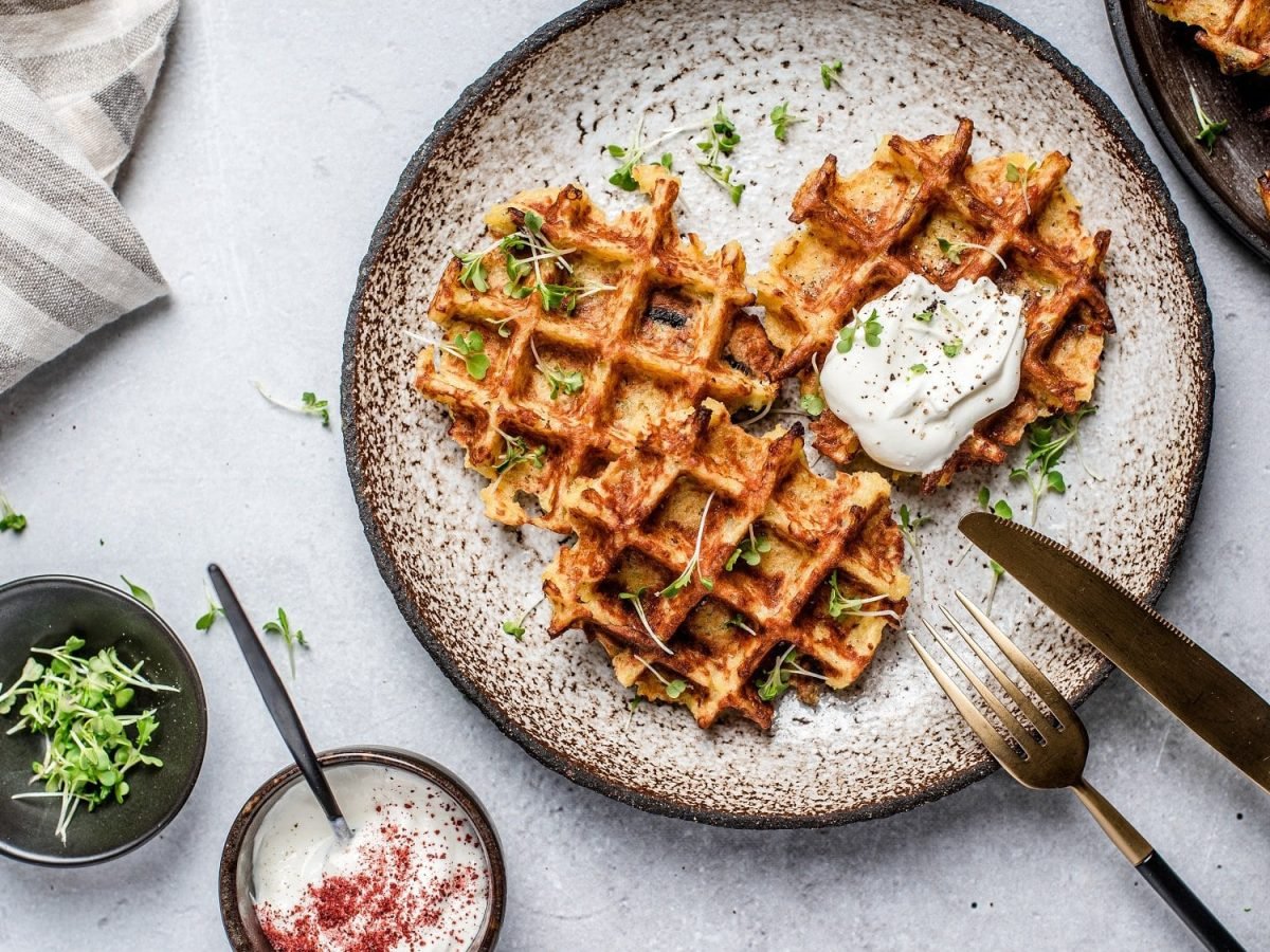 Ein Teller Reiswaffeln aus dem Waffeleisen, die mit einem Klecks Schmand bedeckt sind, daneben Messer und Gabel, ein Geschirrtuch, eine Schale mit Kräutern und Dip und ein Teller weiterer Waffeln.