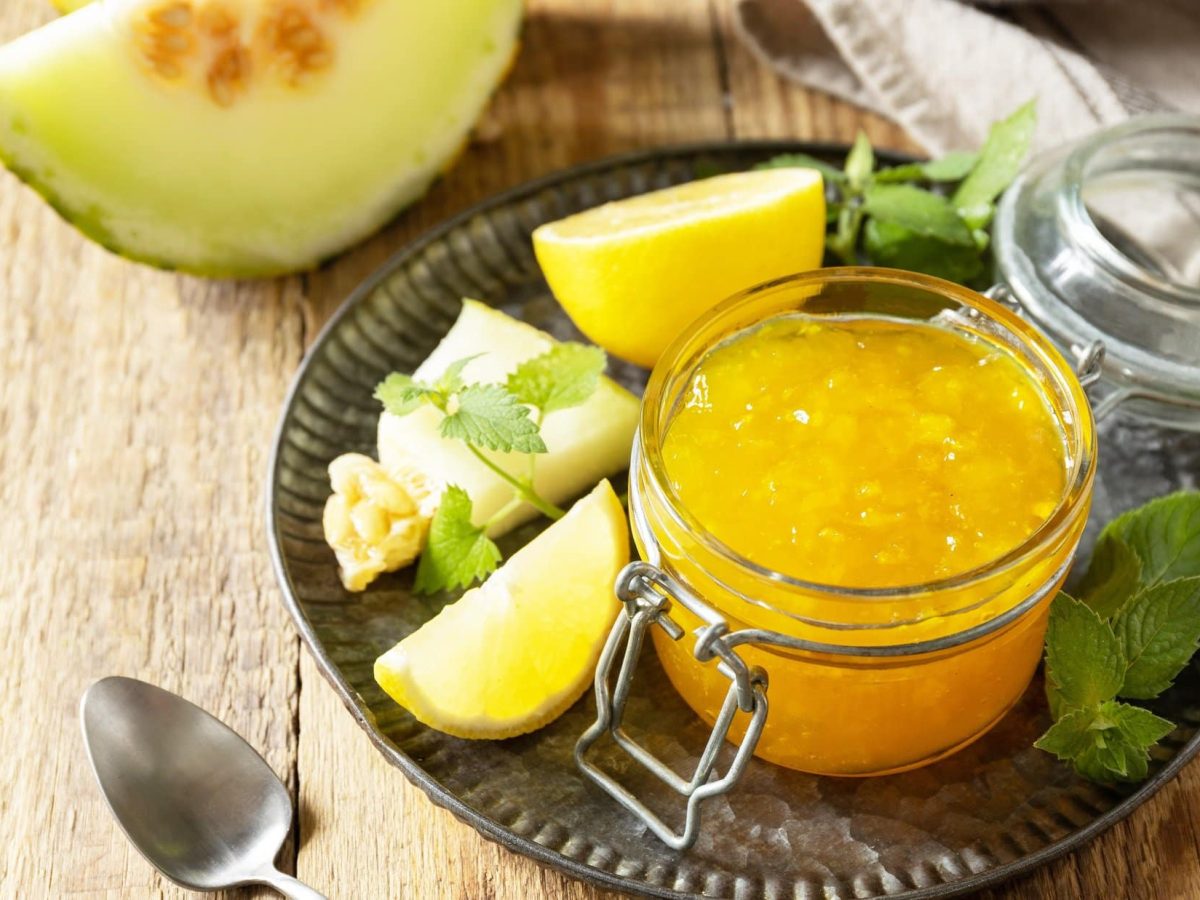 Draufsicht: ein Glas Honigmelonen-Marmelade auf einem dunklen Tablett. Drum herum liegen Honigmelonen-Stücke und frische Kräuter.