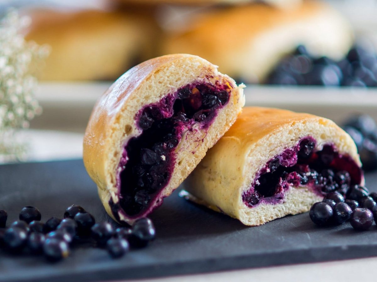 zwei angeschnittene Jagodzianki, polnische Hefebrötchen mit Blaubeerfüllung, auf einer Schieferplatte, drumherum weiße Blüten und frische Blaubeeren.