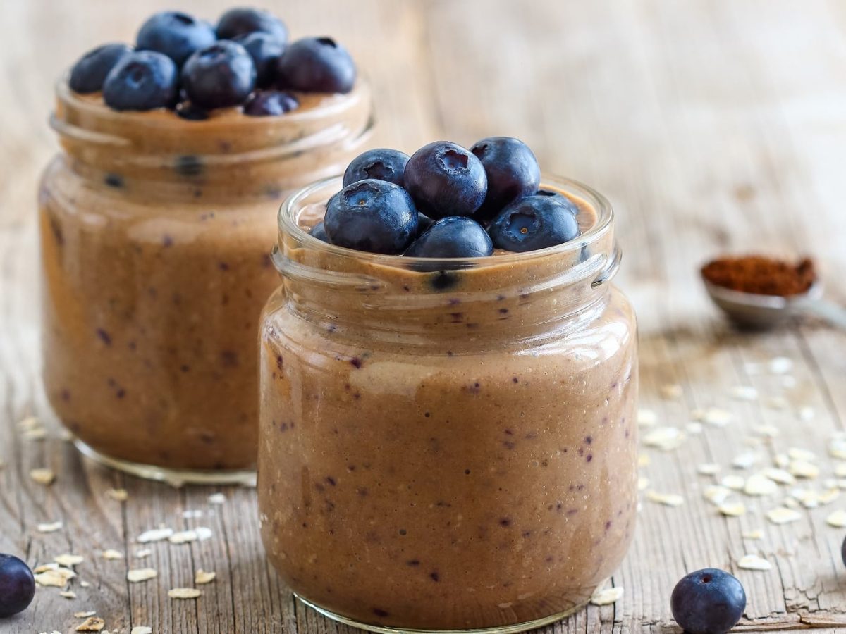 Zwei Gläser mit dem Kaffee-Smoothie und blaubeeren auf einem hellen Holztisch mit Haferflocken seitlich fotografiert.