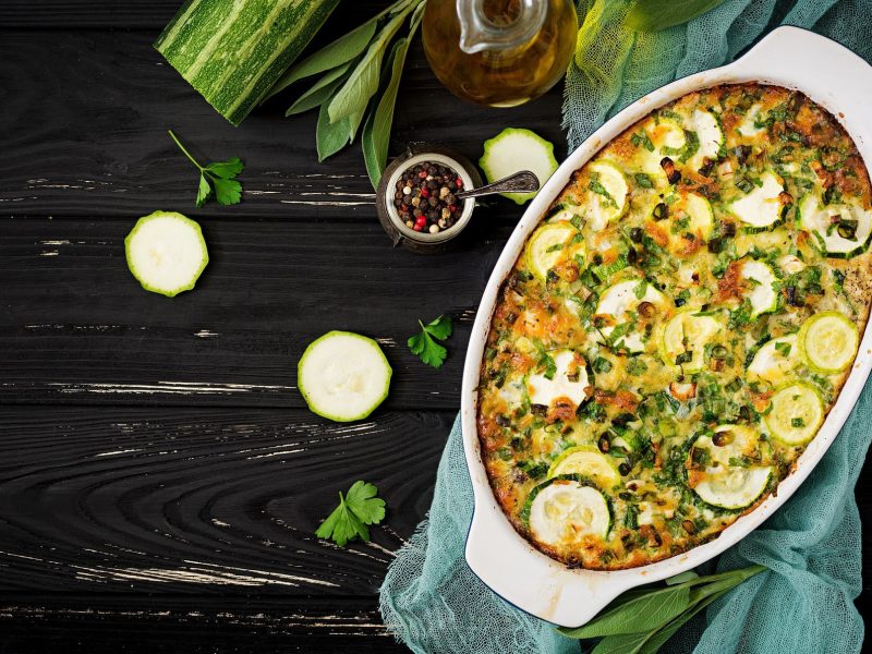 Kartoffel-Lauch-Auflauf mit Zucchini in weißer Auflaufform auf dunklem Holz. Daneben liegen verschiedene Zutaten.