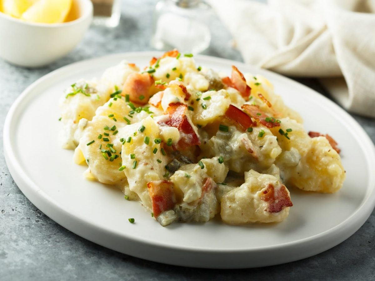 Ein weißer Teller mit dem Kartoffelsalat mit Speck und Cornichons auf grauem Untergrund. Im Hintergrund eine helle Serviette sowie Salz- und Pfefferstreuer.