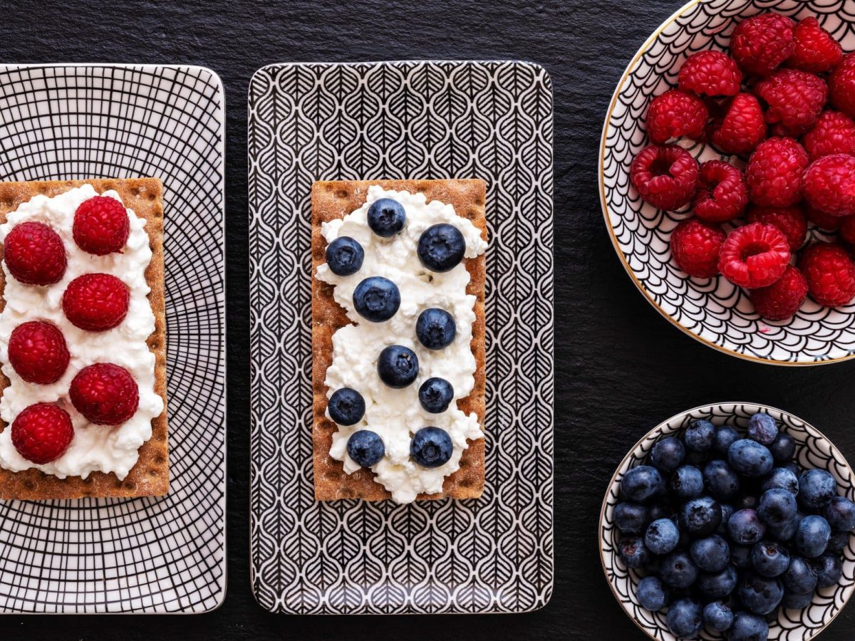 Zweimal Knäckebrot mit frischen Beeren und Vanillesaufstrich auf Frühstücksplatte auf dunklem Untergrund. Daneben zwei Schalen mit Beeren. Draufsicht.