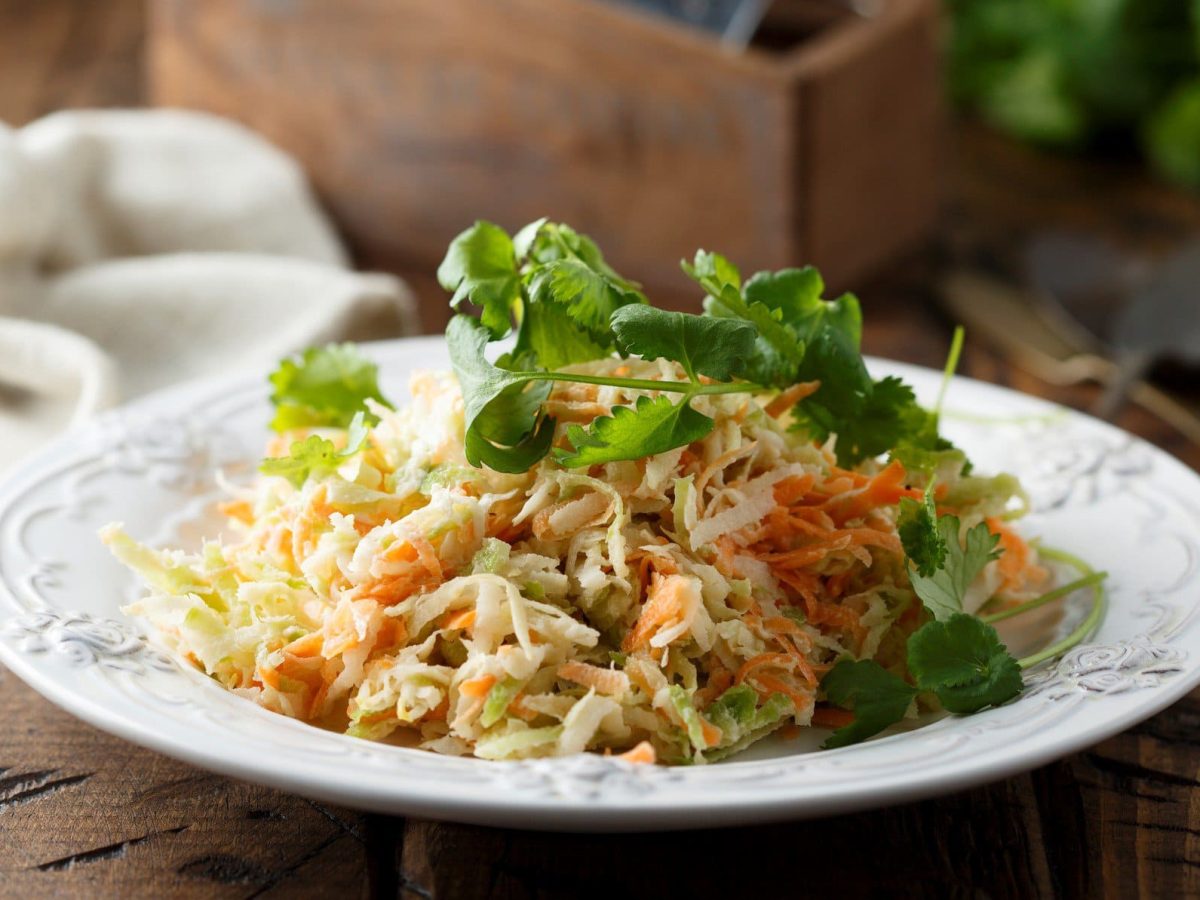 Kohlrabi Slaw mit frischen Kräutern auf weißem Teller und Holzbrett. Im Hintergrund weißes Tuch, eine Holzschachtel und Kräuter. Frontalansicht.