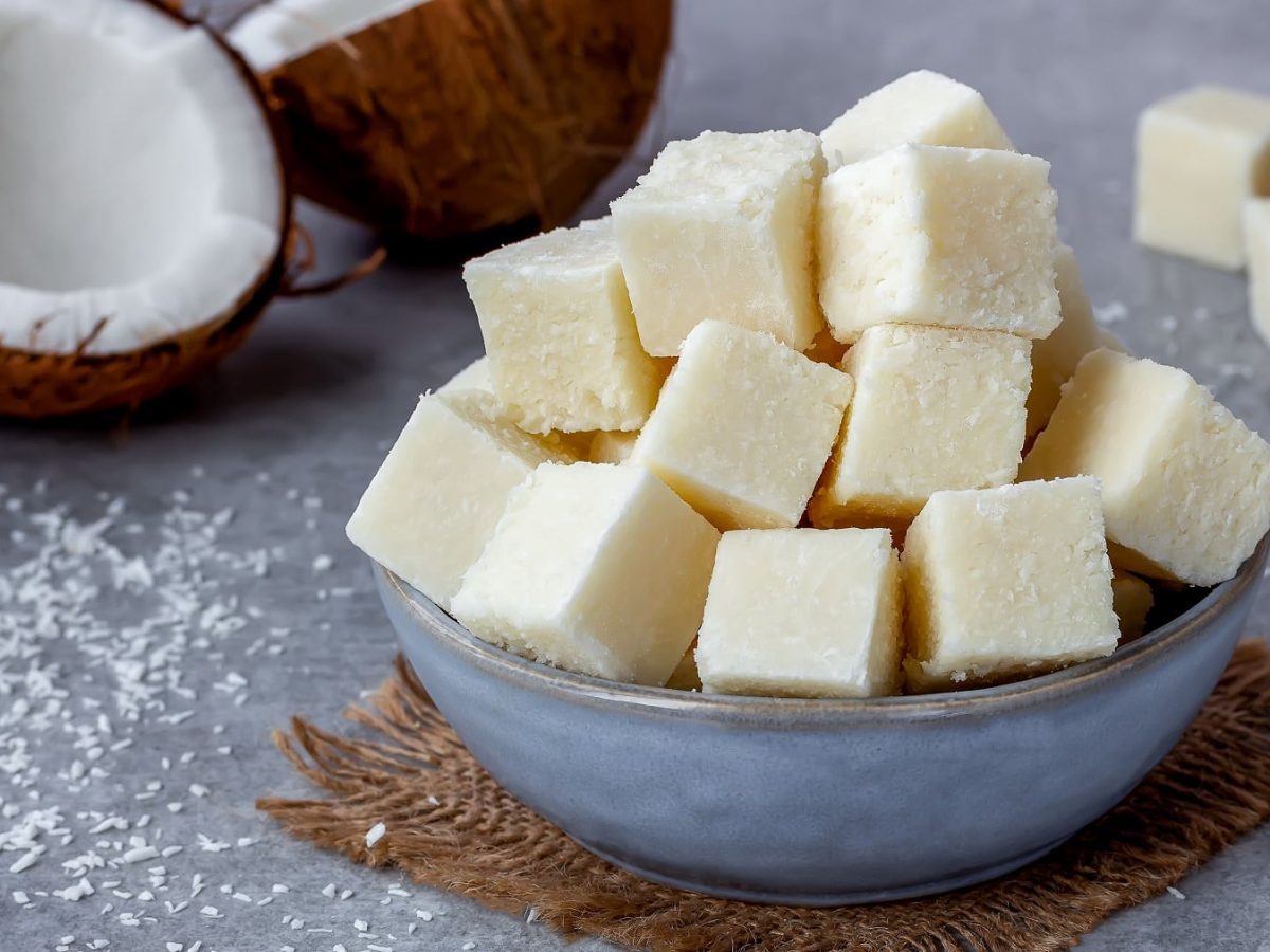 Weiße Kokos-Fudge-Würfel in einer blauen Schüssel auf einem grauejn Tisch mit frischer Kokosnuss und Koksraspel.