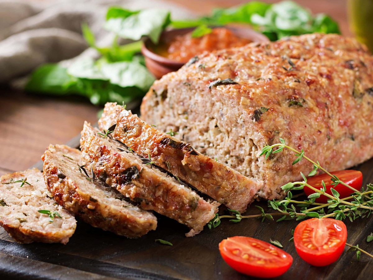 Ein Kräuter-Hackbraten auf einem dunklen Schneidebrett mit Tomaten und Thymian.