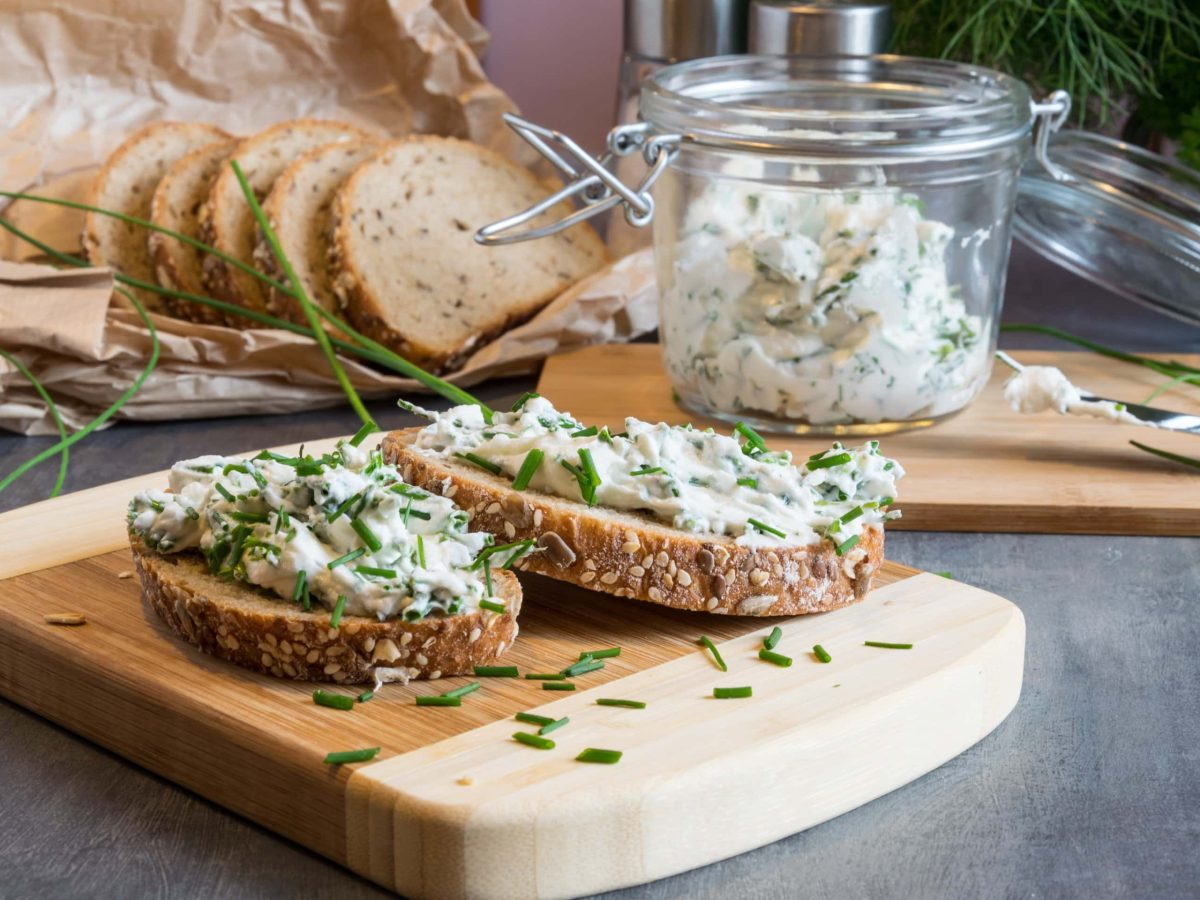 Frontal: Tisch, ein Brettchen mit zwei Scheiben Brot, die mit einem scharfen Kräuter-Hüttenkäse-Aufstrich bestrichen sind. Dahinter ein Glas Scharfer Kräuter-Hüttenkäse-Aufstrich, sowie weitere Brotscheiben. Drum herum liegt frischer Schnittlauch.