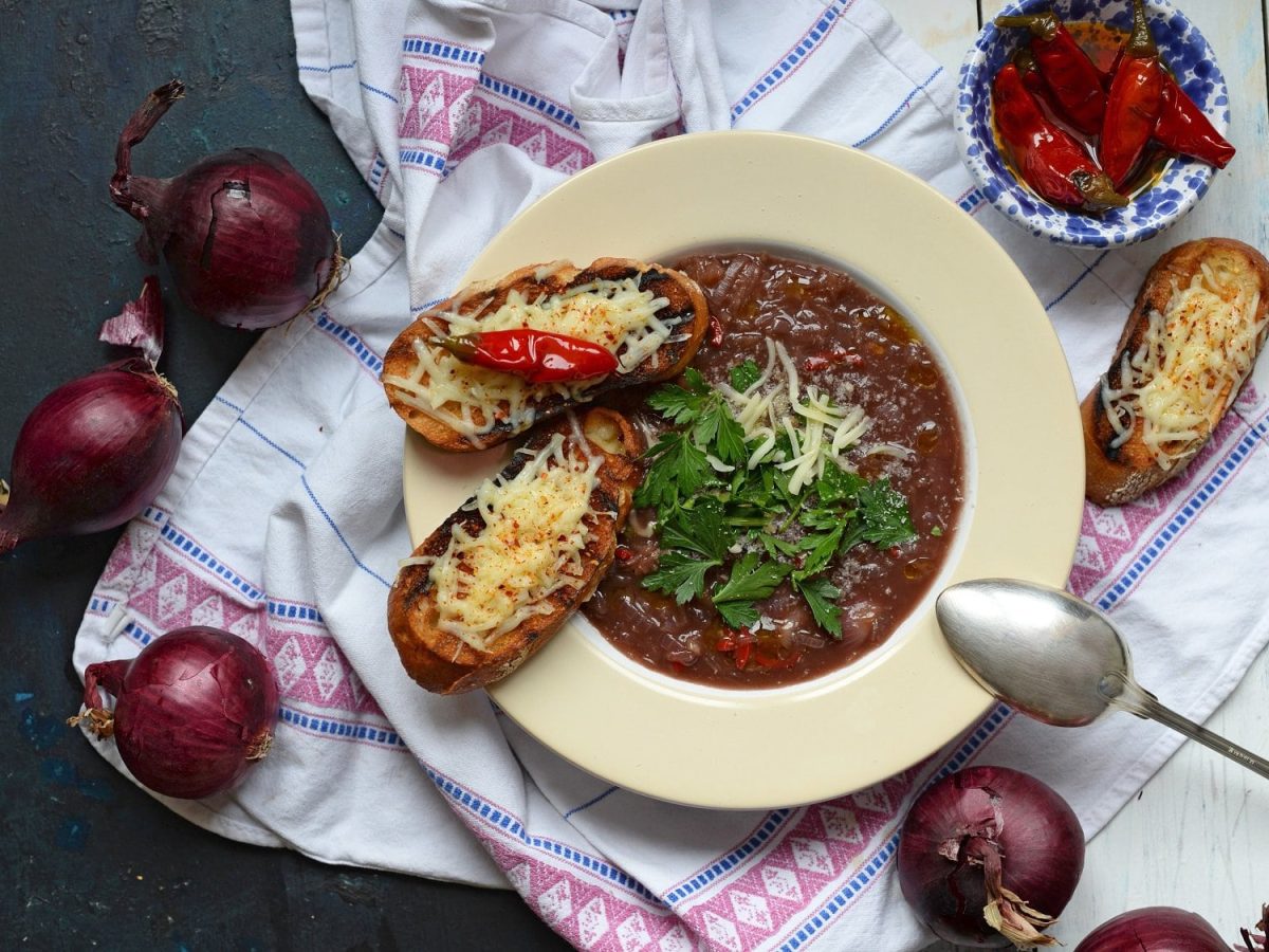 Ein Teller italienische Zwiebelsuppe Licurdia in der Draufsicht, darauf ein Löffel und geröstetes Weißbrot, daneben rote Zwiebeln.