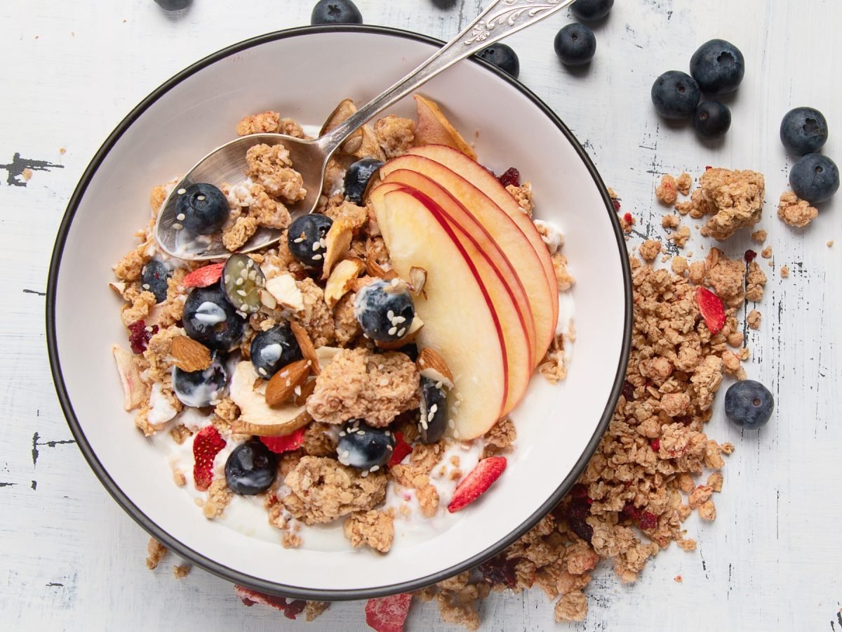 Müsli selber machen mit Joghurt, Blaubeeren, Apfelscheiben, Mandeln und Sesam in einer weißen Schale mit Löffel.