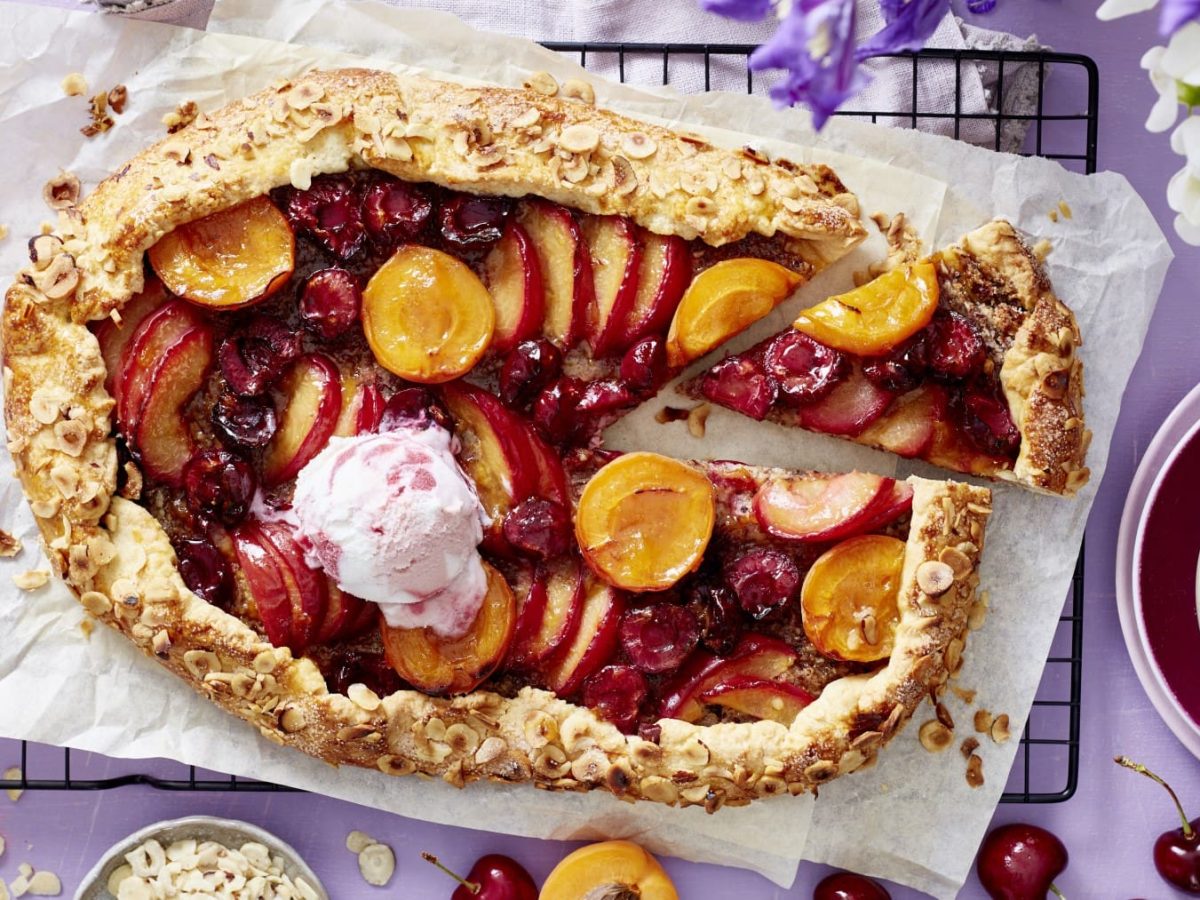 Eine angeschnittene Obst-Galette mit einer Kugel Eiscreme auf Backpapier, darunter ein schwarzes Gitter auf lila Untergrund, drum herum Früchte und Geschirr.