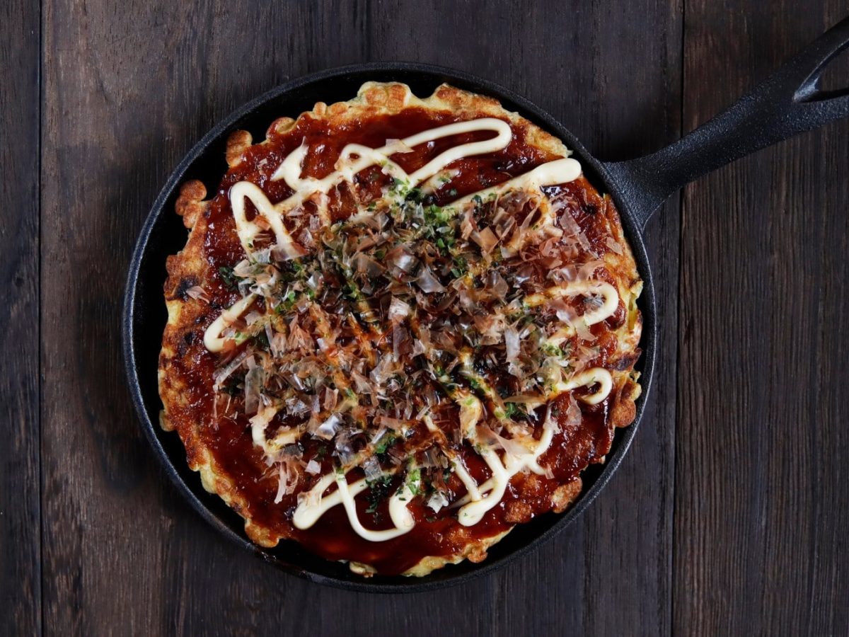 Eine schwarze Pfanne mit Okonomiyaki von oben fotografiert vor einem hölzernen Hintergrund.