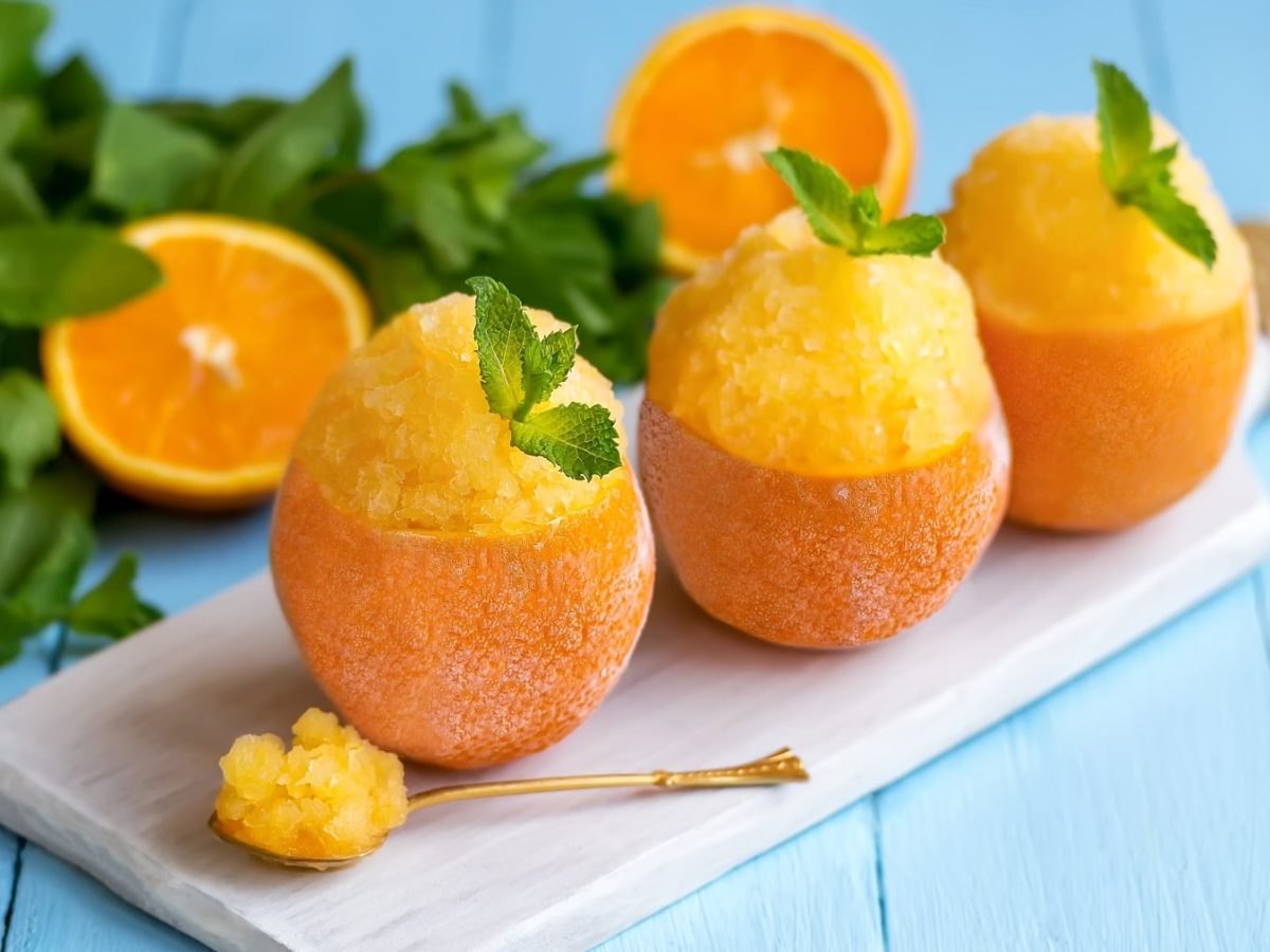 Drei ausgehöhlte Orangen mit dem Granita auf einem kleinen Schneidebrett und hellblauem Holztisch. Im Hintergrund Minze und Orangenhälften.