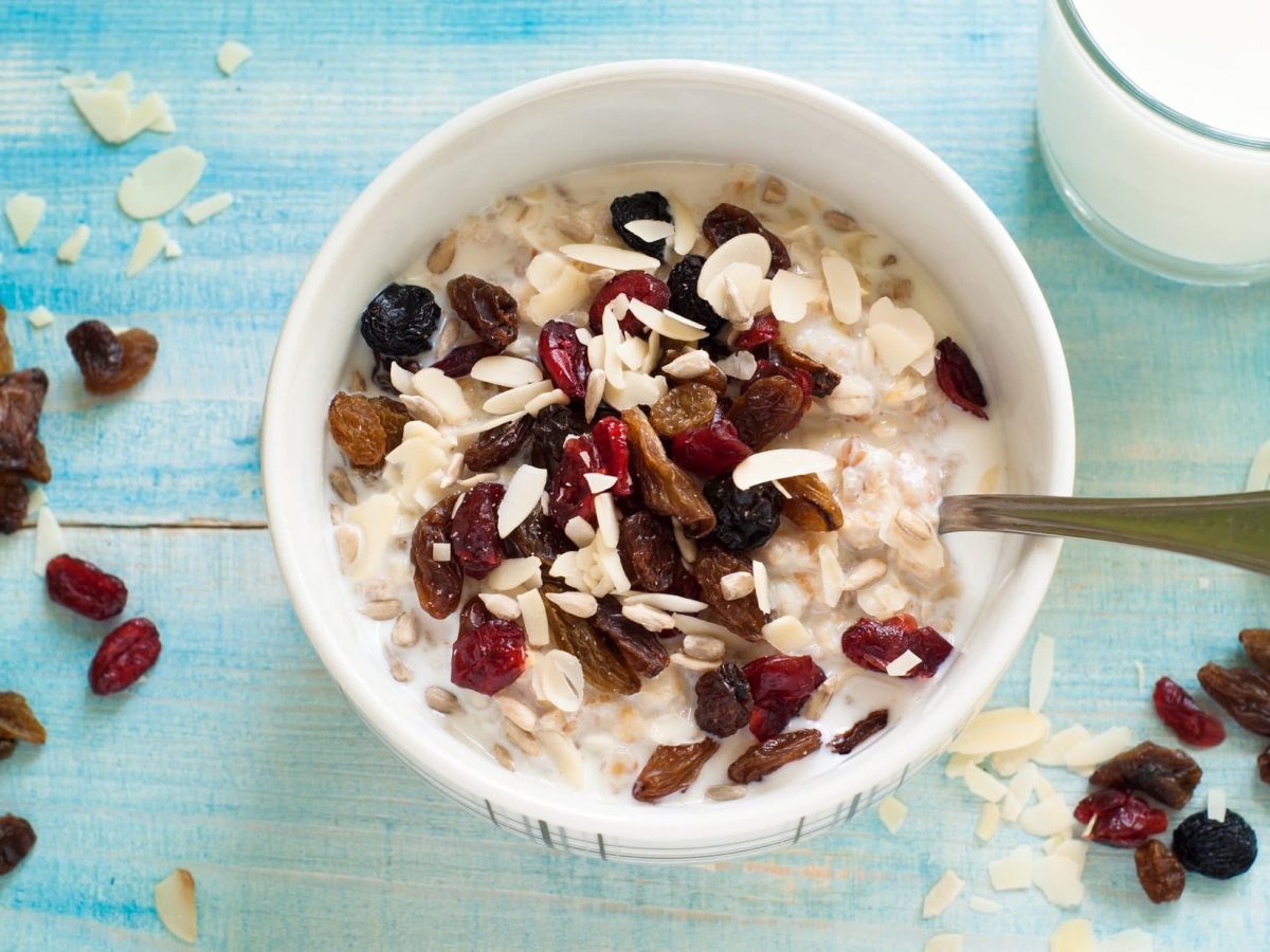 Hellblauer, melierter Untergrund aus Holz: Eine Bowl Overnight-Oats mit getrockneten Früchten und Mandeln steht darauf. Daneben steht ein Glas Milch. Auch Mandeln und getrocknete Früchte sind verstreut.