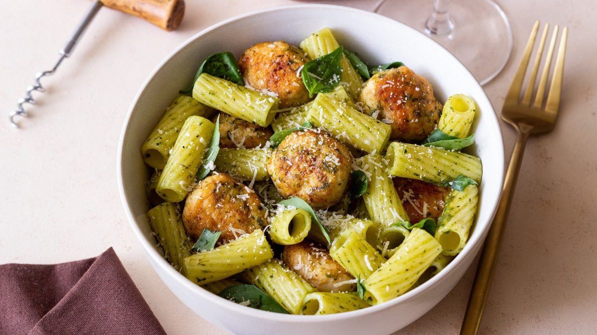 Eine weiße Schale mit der Pesto-Pasta und den Hackbällchen auf einem hellen Tisch mit brauner Tischdecke und einer goldenen Gabel.