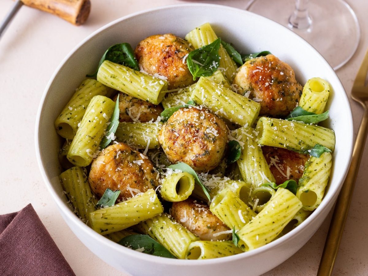 Eine weiße Schale mit der Pesto-Pasta und den Hackbällchen auf einem hellen Tisch mit brauner Tischdecke und einer goldenen Gabel.