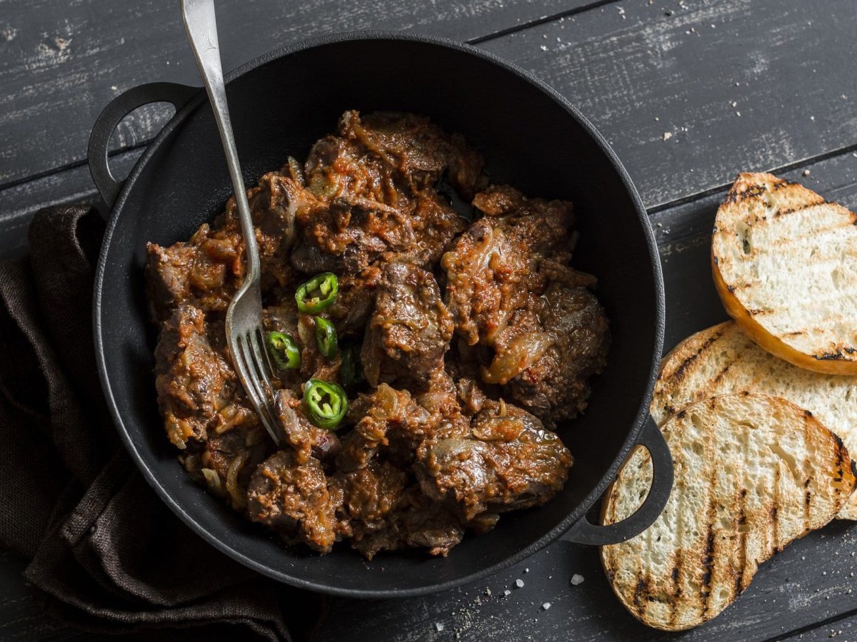 Draufsicht: Dunkler Untergrund aus Holz, eine dunkler offener Topf mit Piri-Piri-Hühnerleber und daneben liegen zwei geröstete Weißbrotscheiben.