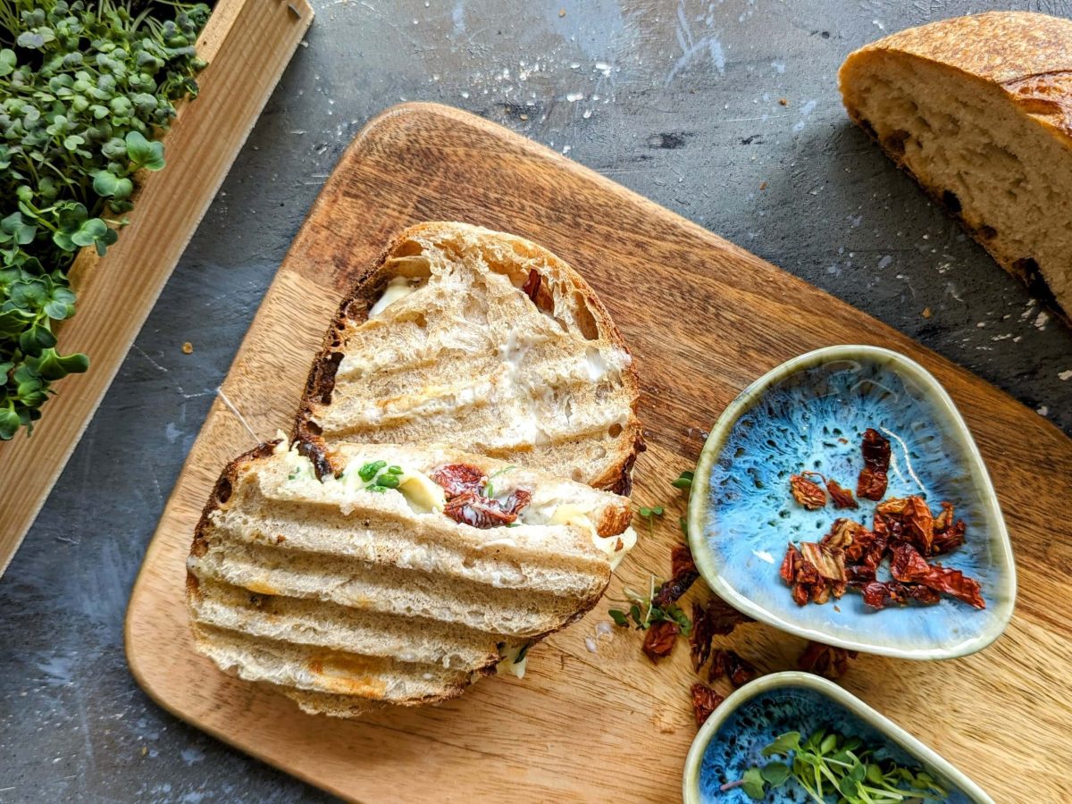 Das halbierte Sandwich auf einem Holzbrett. Daneben Kresse, Brot und getrocknete Tomaten.