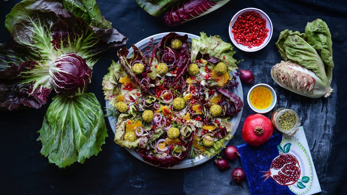 Radicchio mit Pistazien-Ricotta-Bällchen, Orangen und Granatapfelkernen in der Draufsicht, dunkelblauer Untergrund, daneben viele zutaten wie Salat und Granatapfel.